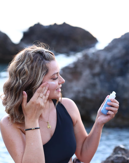 A woman holding a bottle of mastic daytime moisturizer with the blue waters of a Greek pebble beach as the backdrop